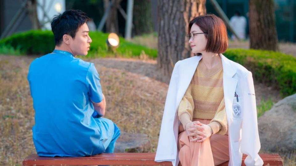 A man in scrubs sits on a bench with his back to the camera with a woman in a doctor's coat sits next to him, facing forward and smiling at him