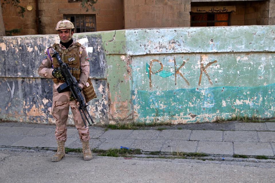 Iraqi army soldier stands next to the graffiti left by an affiliate of the Kurdistan Worker's Party, or PKK, which recently withdrew in Sinjar, Iraq. Friday Dec. 4, 2020. A new agreement aims to bring order to Iraq's northern region of Sinjar, home to the Yazidi religious minority brutalized by the Islamic State group. Since IS's fall, a tangled web of militia forces have run the area, near the Syrian border. Now their flags are coming down, and the Iraqi military has deployed in Sinjar for the first time in nearly 20 years. (AP Photo: Samya Kullab)