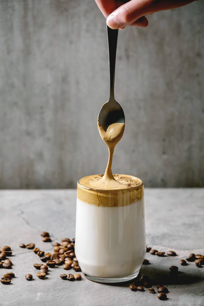 Person's hand drizzling whipped coffee over milk in a glass, surrounded by scattered coffee beans