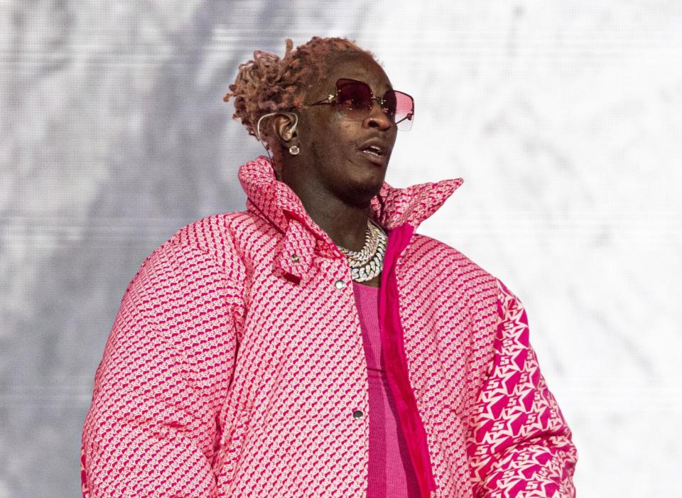 Young Thug stands onstage while wearing sunglasses, gold chains and a pink patterned puffer jacket