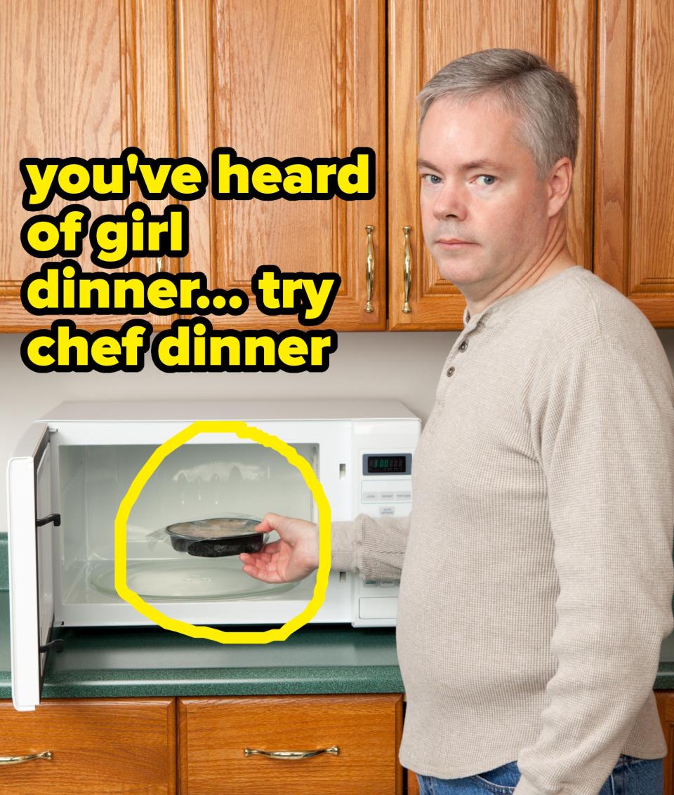 A man with gray hair and a light sweater holds a pan while using a microwave in a kitchen with wooden cabinets