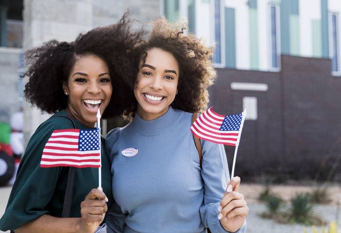 Two women smile while holding American flags. Both women are wearing "I Voted" stickers