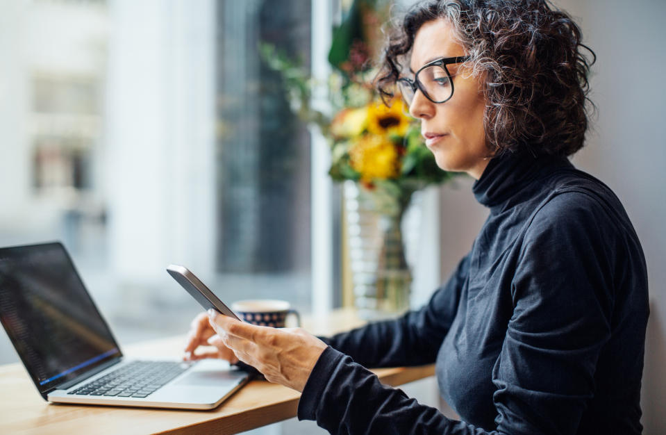 Woman in glasses using smartphone and laptop at desk, with flowers in background