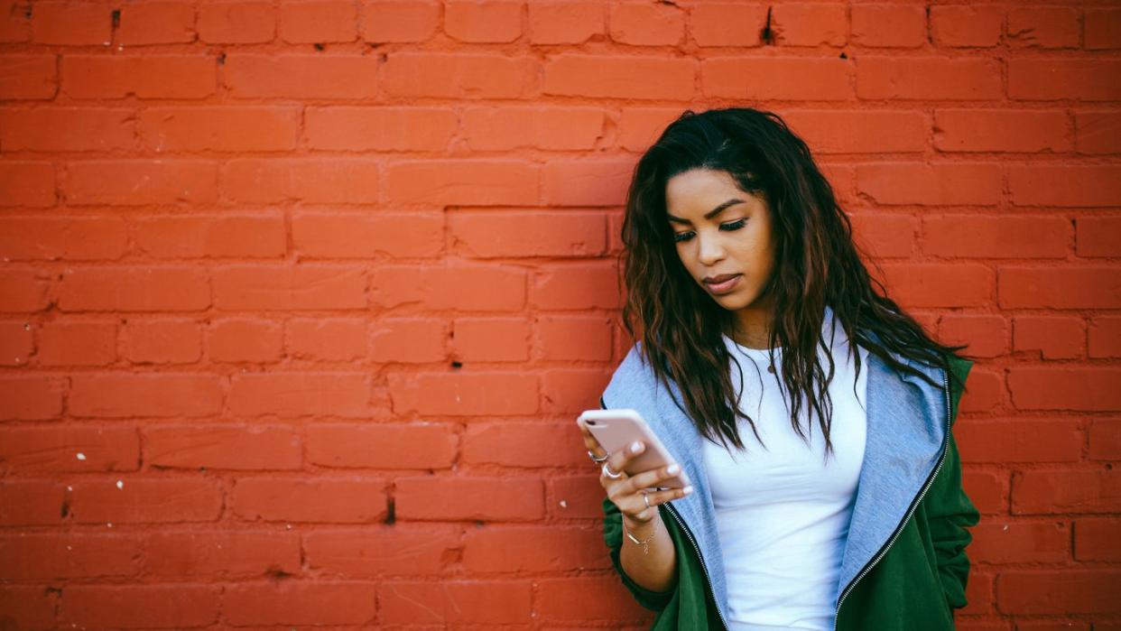 woman looking pensively at smartphone against brick wall