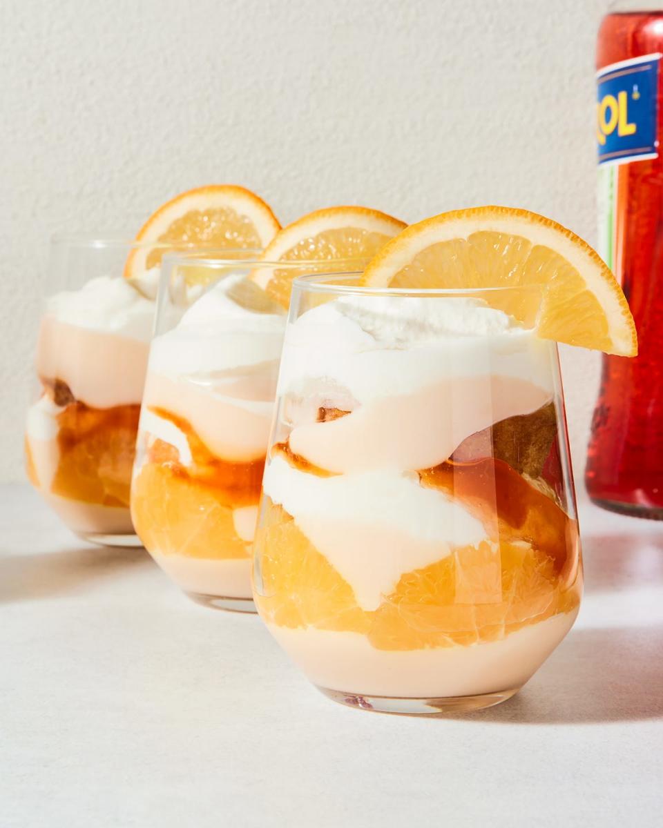 a stemless wine glass filled with vanilla cake, whipped cream, aperol spiked jam, and pudding with an orange slice on the rim