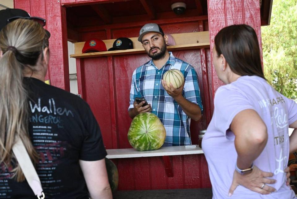 Erik Alfaro, de Del Bosque Farms, en el centro, muestra dos melones a los compradores en el puesto de frutas de la familia en Shields Avenue, cerca de la I-5 en el extremo oeste del Valle, el lunes 15 de julio de 2024.