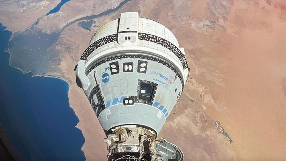 a white space capsule is seen docked to the International Space Station, with a coastal desert landscape below