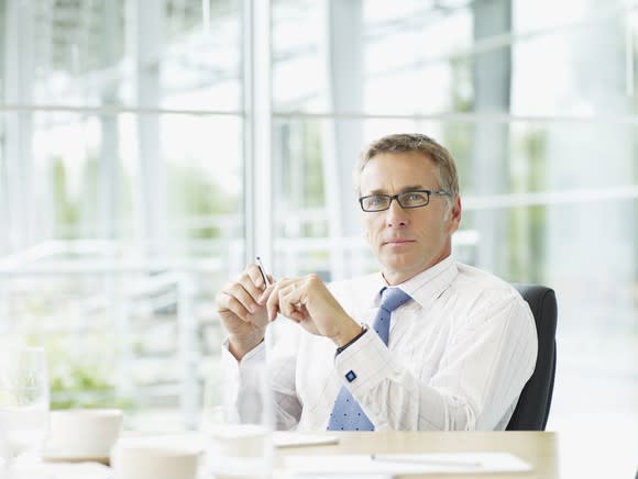 A businessman in a dress shirt and tie sitting behind his desk in front of a row of glass windows.