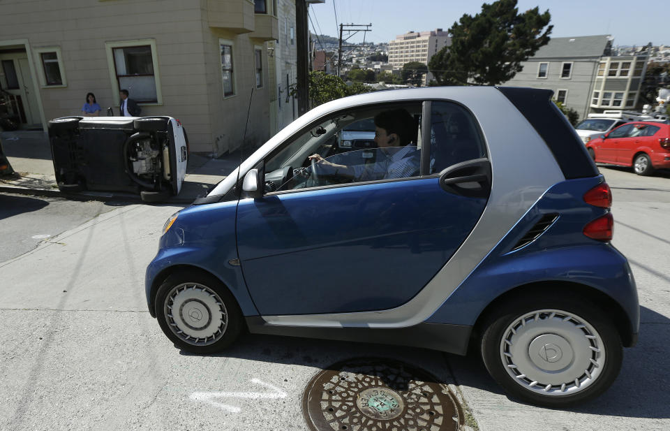 CORRECTS TO PROSPECT AND COSO AVENUES- A woman in a Smart car drives past a tipped over Smart car on the corner of Prospect and Coso Avenues in San Francisco, Monday, April 7, 2014. Police in San Francisco are investigating why four Smart cars were flipped over during an apparent early morning vandalism spree. Officer Gordon Shyy, a police spokesman, says the first car was found flipped on its roof and a second was spotted on its side around 1 a.m. Monday in the Bernal Heights neighborhood. (AP Photo/Jeff Chiu)