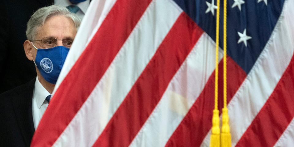 Merrick Garland wears a mask standing behind the american flag