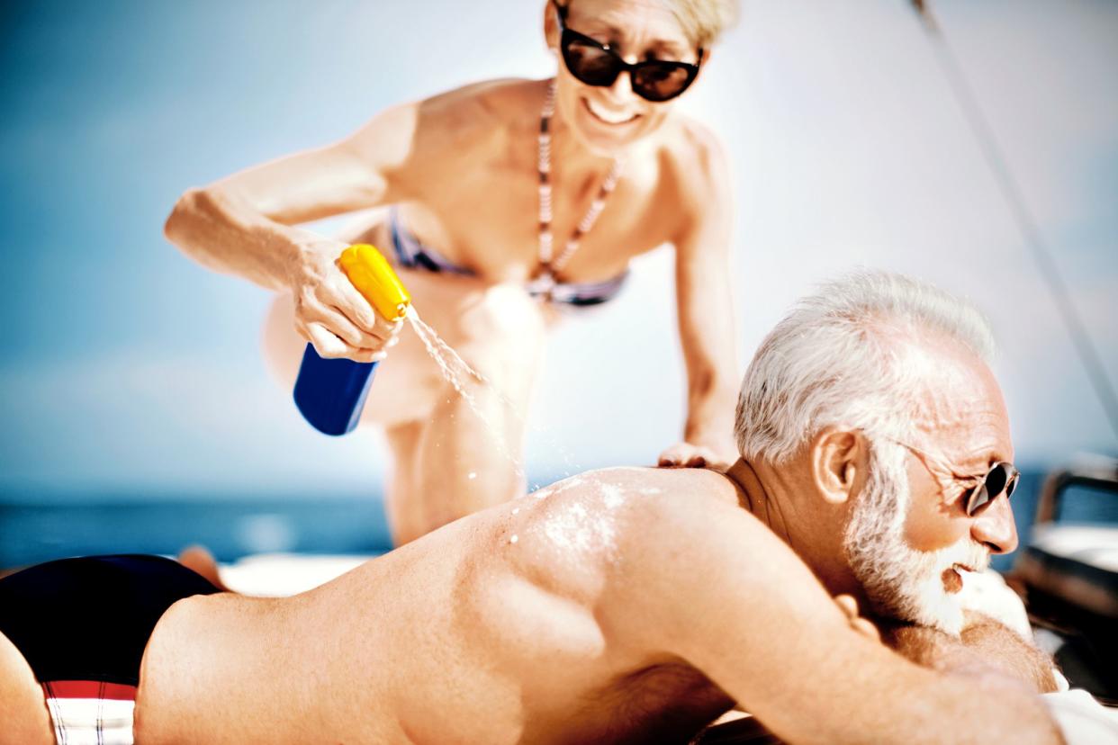 Senior woman applying sunscreen at the back of her husband while he's lying on the stomach and enjoying at yacht.