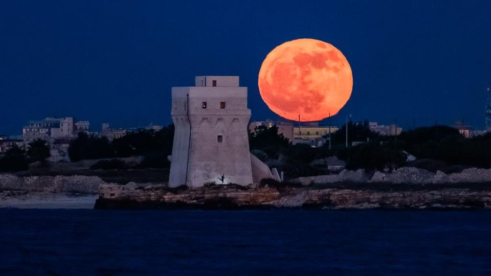 there is a large red orange moon in the background.  A white stone tower in the foreground with the silhouette of a dancer visible in front of it.