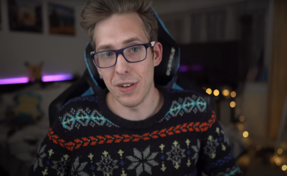 Evan Edinger is sitting in a room with a gaming chair, wearing glasses and a patterned sweater. The room has a cozy setup with string lights in the background