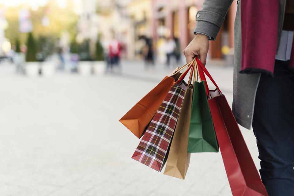 Hand holding multiple shopping bags, with outdoor retail shops blurred in the background.