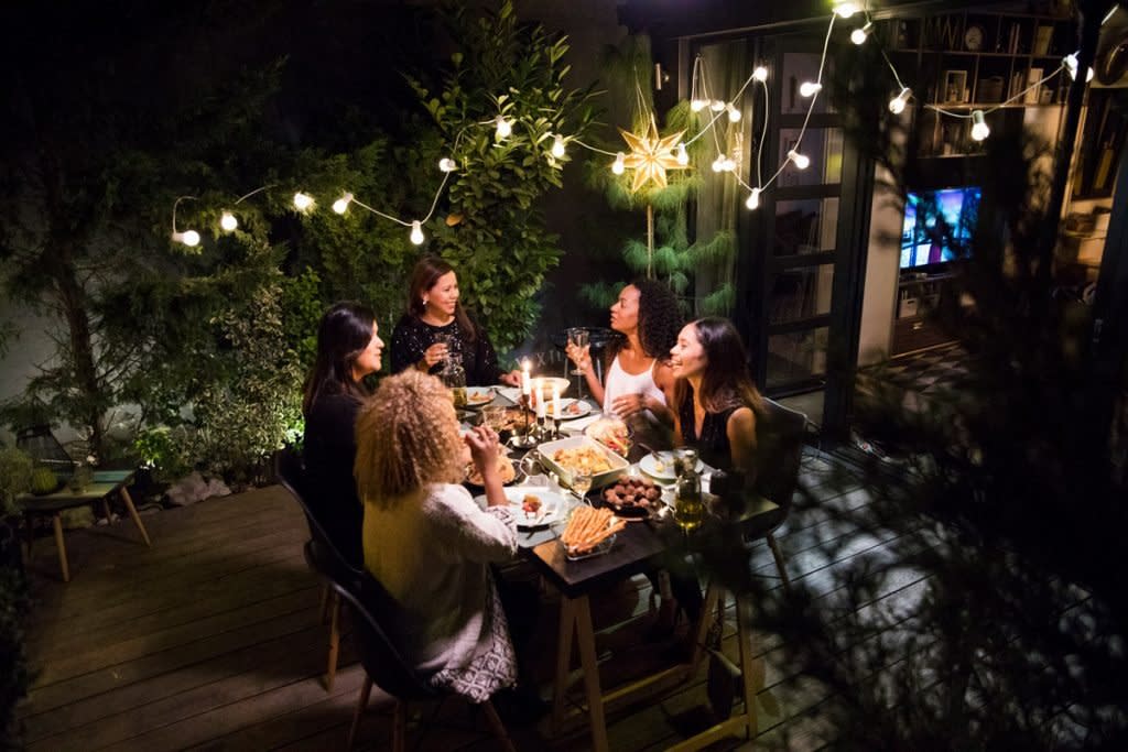 A group of women enjoy dinner outside on someone's back patio with lights strung up all around them.