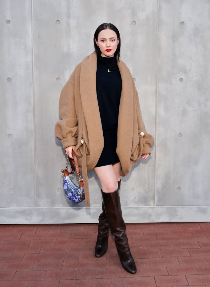 Iris Apatow wearing a black mini dress with a camel coat and dark brown boots. - Credit: Louis Vuitton