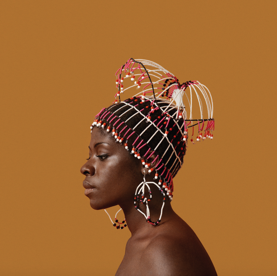 An image from Kwame Brathwaite's "Black is Beautiful," features a Black woman's side profile with bare shoulders wearing an intricate beaded headpiece in white, pink and red.