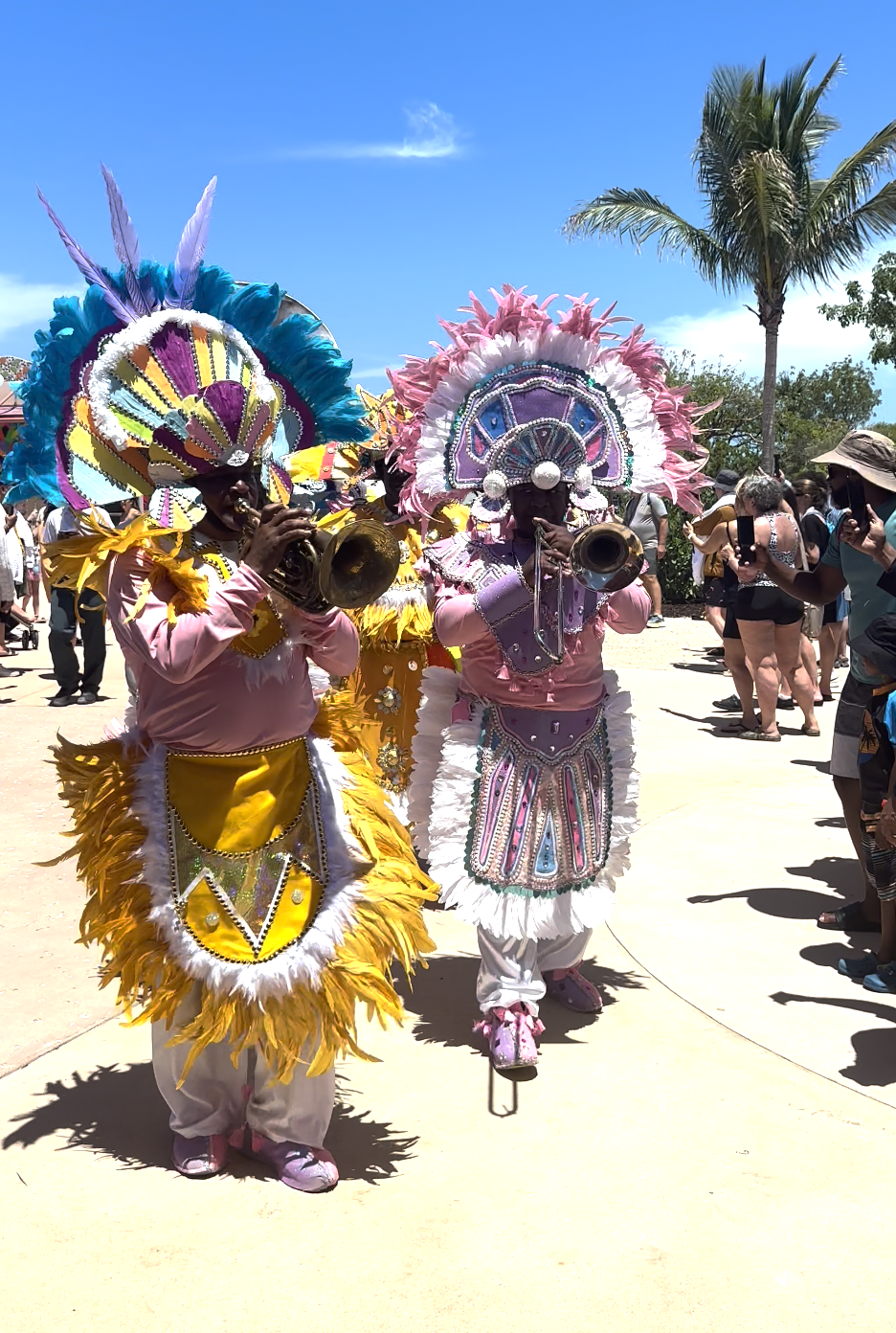 Two performers in elaborate feathered headdresses and decorated costumes play trumpets at an outdoor event. They're surrounded by an audience