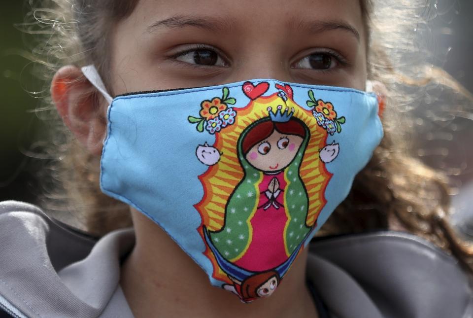 A Venezuelan migrant girl, wearing a protective face mask with a graphic version of the Virgin of Guadalupe, waits for a bus to travel to the border, after breaking camp in Bogota, Colombia, Thursday, July 2, 2020. Facing no work due to the COVID-19-related economic shutdown, hundreds of Venezuelan migrants are returning to their country. (AP Photo/Fernando Vergara)