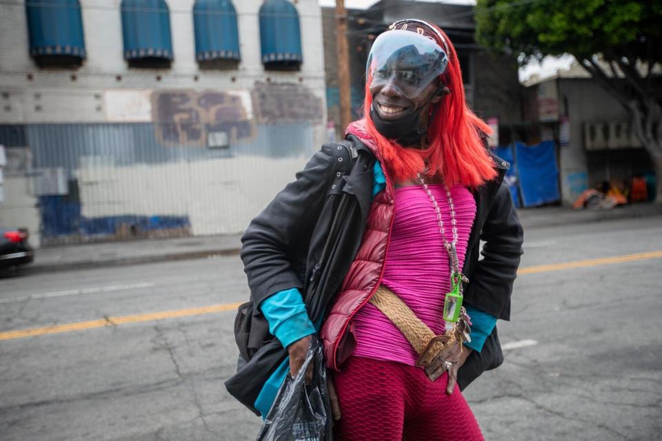 A person in brightly colored clothing and dyed hair smiles with hands on hips.