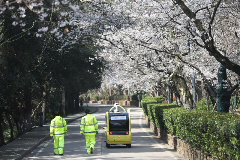 Security personnel walk next to a 5G enabled autonomous vehicle, installed with a camera filming blooming cherry blossoms for an online live-streaming session, inside the closed Wuhan University