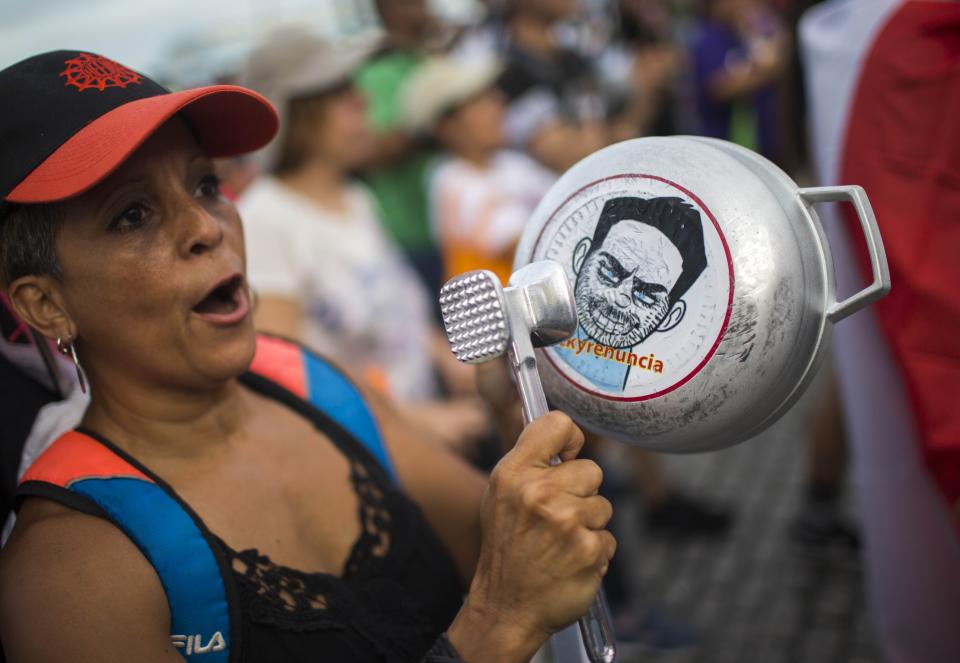 FILE - In this July 19, 2019 file photo, a demonstrator bangs on a pot that has a cartoon drawing of Governor Ricardo Rossello and text the reads in Spanish "Quit Ricky" as people gather to protest against Gov. Rossello, in San Juan, Puerto Rico. Puerto Ricans are now asking themselves what comes after Gov. Ricardo Rossello announced his resignation after having achieved their main goal: ousting a governor for the first time in the U.S. territory's recent history. (AP Photo/ Dennis M. Rivera Pichardo, File)