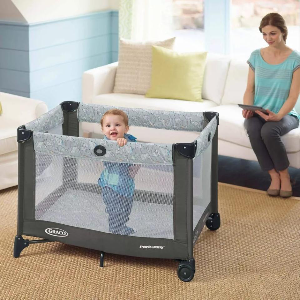 Toddler model standing inside a gray and black pack and play with mesh sides