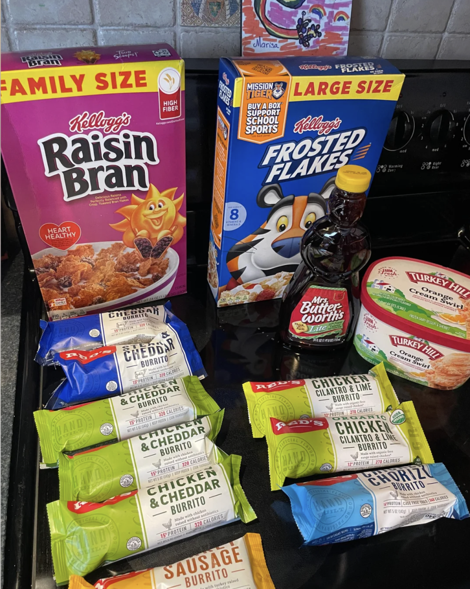 Various family-size breakfast cereals, syrup, frozen turkey meals, and an assortment of burritos are displayed on a kitchen counter