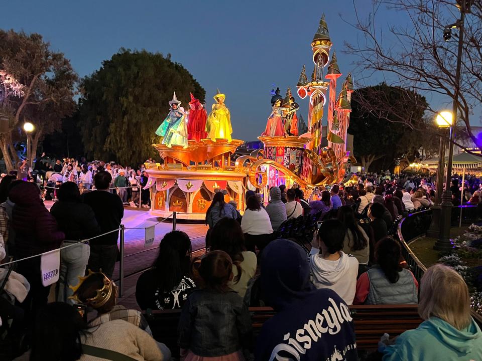 A Disneyland parade at night, where people are seated on benches and in roped-off areas. A float passes by with Sleeping Beauty characters on board.