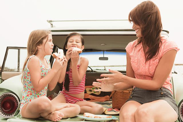 Image Source / Getty Images Mother and daughters eating sandwiches on car boot