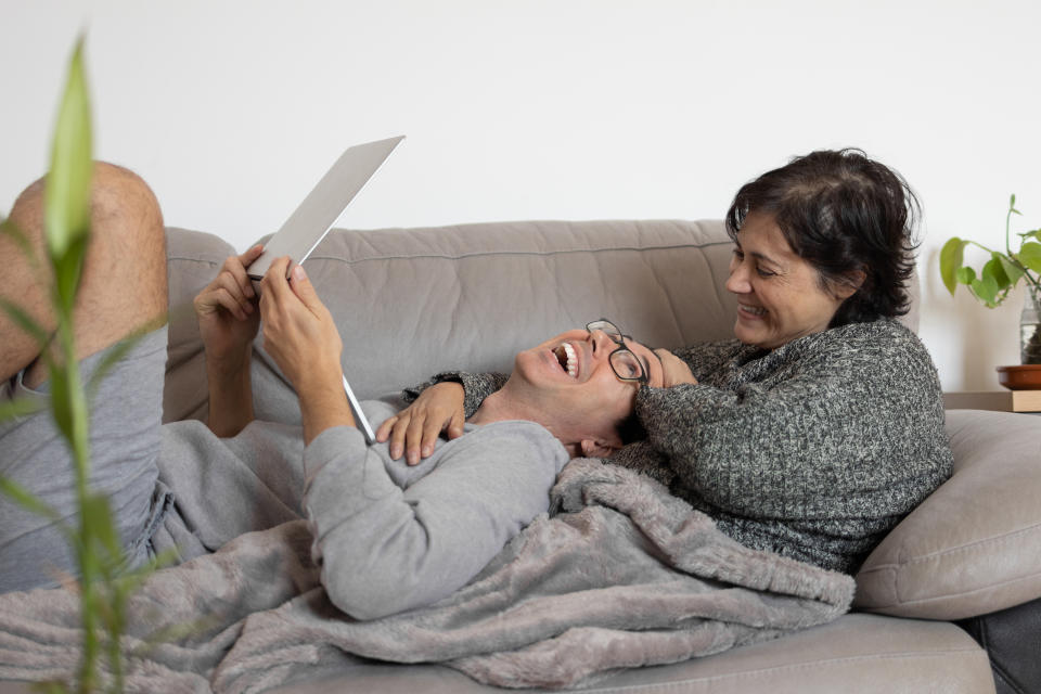 A mature woman with her younger partner covered with a warm blanket on the sofa at home and using a laptop.