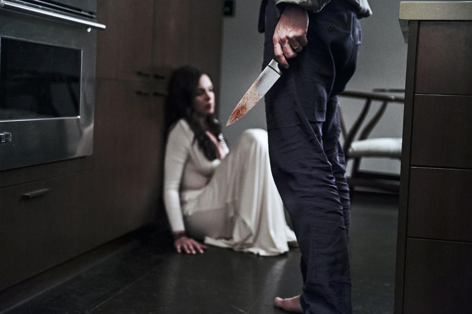 A terrified woman in a white dress crouches near a barefoot man with a kitchen knife