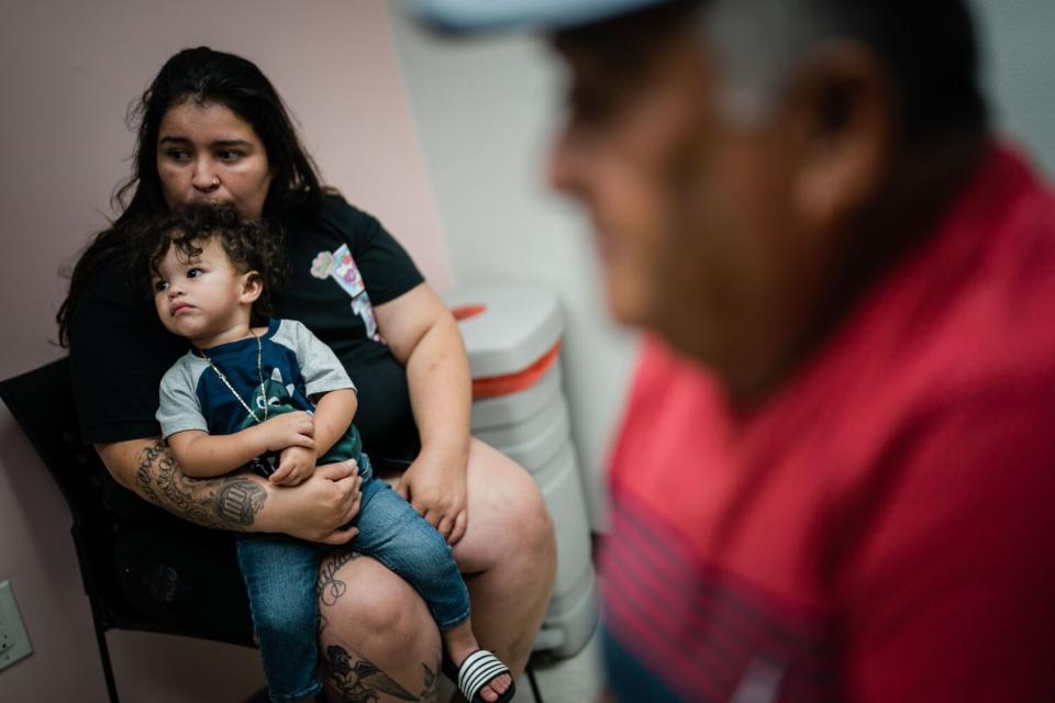 At left a woman holds a baby on her lap while a man sits at right, blurred in the foreground.