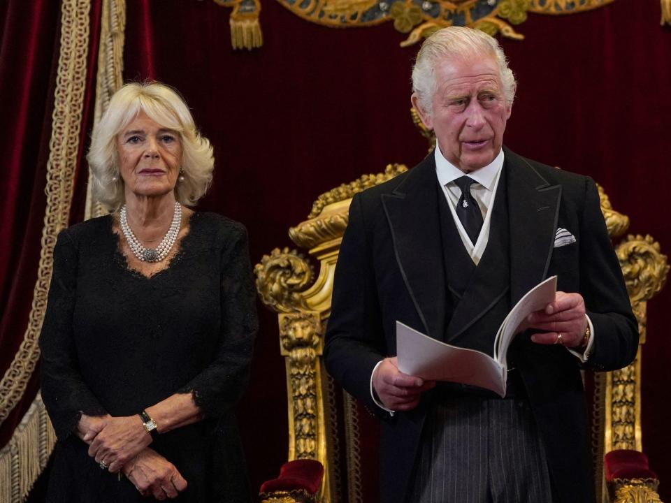 Camilla, Queen Consort stands next to King Charles as he reads from a booklet.