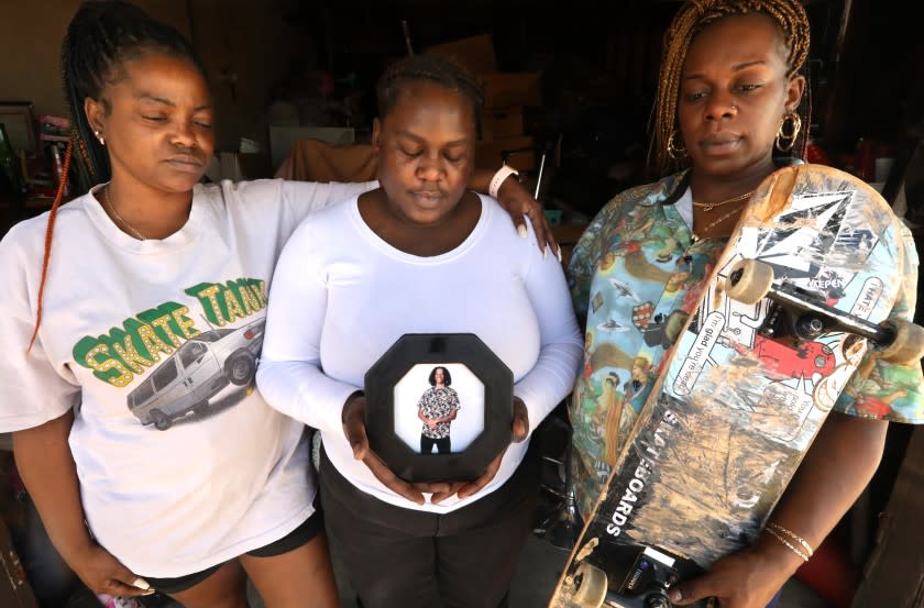 LANCASTER, CA - JUNE 29, 2020 - - Sisters Candice Russaw, 33, from left, Chanel Evans, 28, and Brandy Russaw, 34, remember their brother, 20-year-old Jacob Russaw, in photo, who committed suicide in 2018. Candice wears Jacob's t-shirt and Brandy holds his skateboard and wears one of his shirts. The sisters were photographed at Brandy's home in Lancaster on June 29,. 2020. He was a skilled skateboarder with hopes of going pro or getting a sponsorship. At the time of his death he'd been put out of a youth homeless shelter in Hollywood, Covenant House, for infractions, despite being well-loved by shelter residents and officials, who put on a memorial and paid for his funeral. Candice feels the agency could have done more to provide mental health and prevent his death. Russaw died by hanging himself. (Genaro Molina / Los Angeles Times)