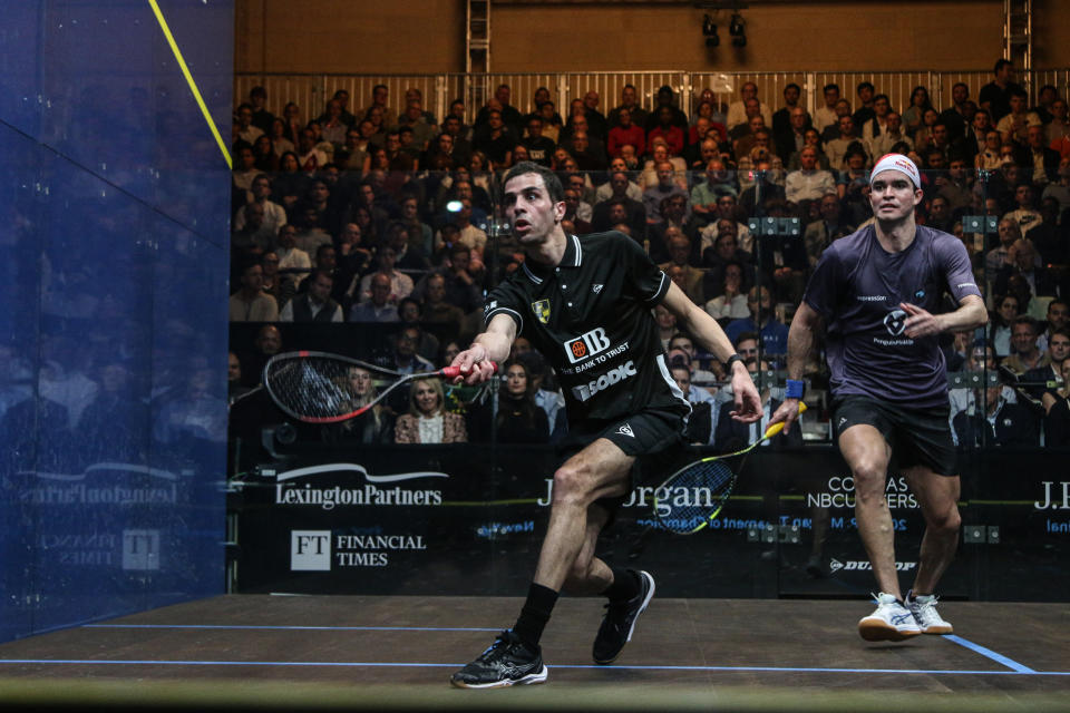 Two men play squash on a professional court while an audience watches behind a glass wall