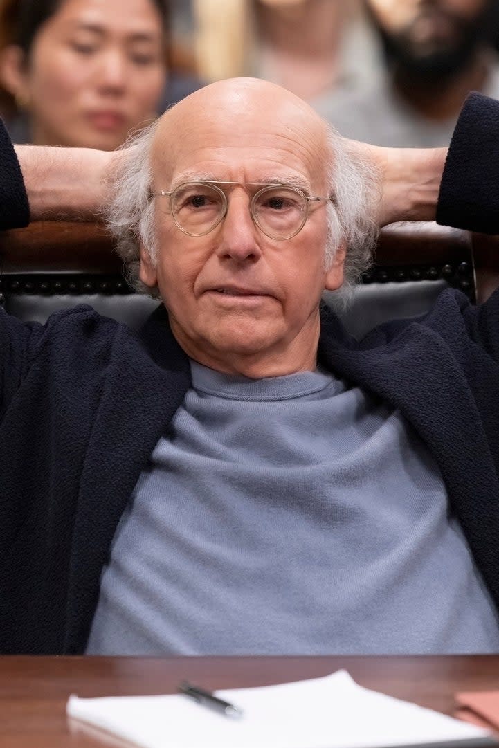 Larry David sits relaxed with hands behind his head at a courtroom setting with people in the background. A notebook and papers are on the table