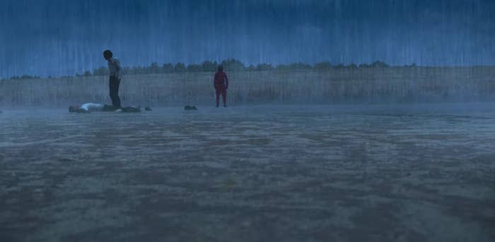 Gi-hun stands over Sang-woo, who lies on the ground in the rain; a guard watches on