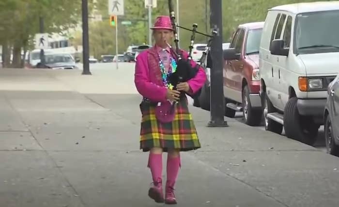 Person in a kilt and hat playing bagpipes on a city sidewalk