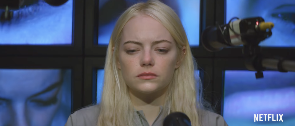 A close up of Emma Stone in Maniac, dressed in grey with screens showing her face on the wall behind her.
