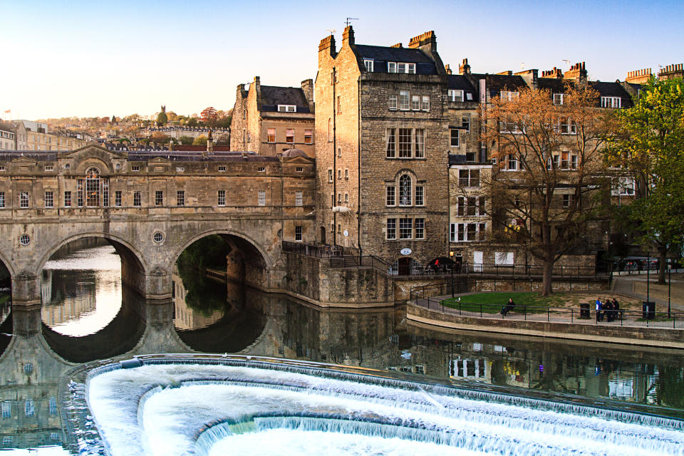 Historic stone buildings by a river with cascading water and a bridge, in a serene urban park setting