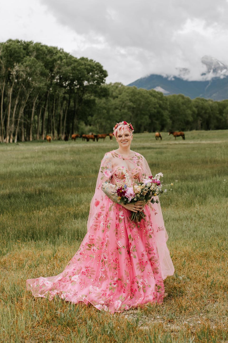 A bride in a pink, floral wedding dress stands in a field with horses.