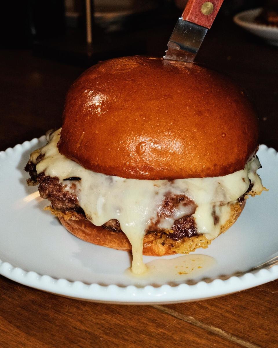 A vertical closeup of a burger dripping with raclette cheese, a knife plunged through the top, at Burgette in Santa Monica