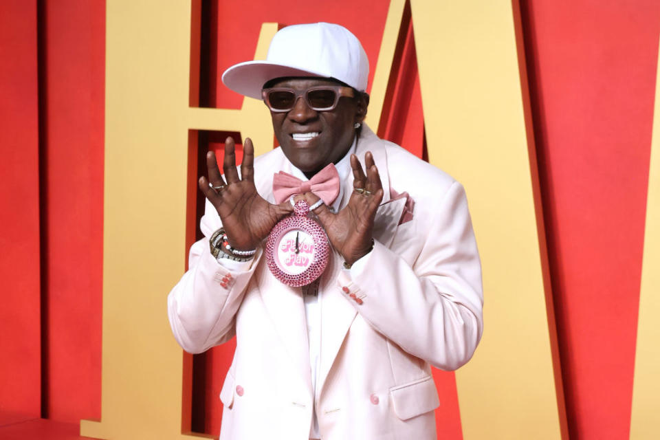 Person posing with hands near face, wearing a wide-brimmed hat, suit with embellished brooch, and large rings