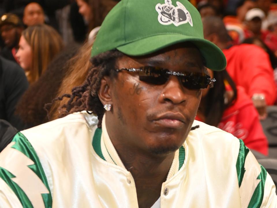 Young Thug sits at a basketball game in a white satin jacket, sunglasses, and green baseball cap.