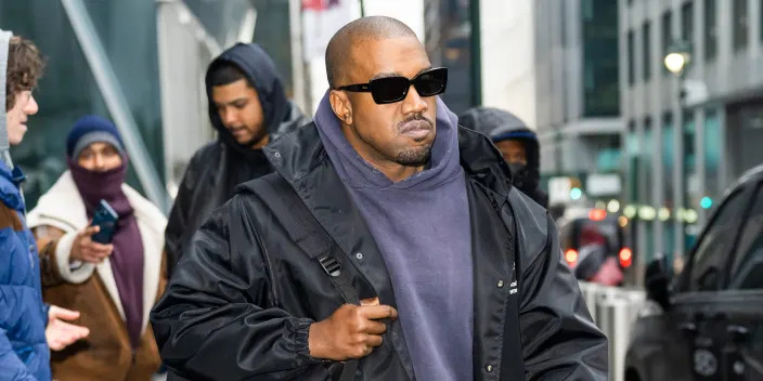 Kanye West walks down a sidewalk with a backpack slung over his shoulder