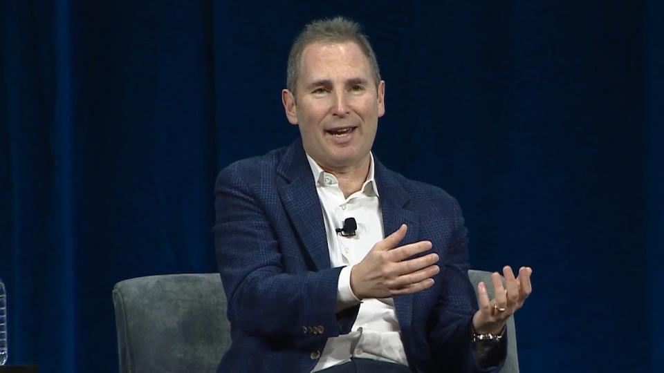 Amazon CEO Andy Jassy gestures to the audience as part of a conversation on stage.  Black background.