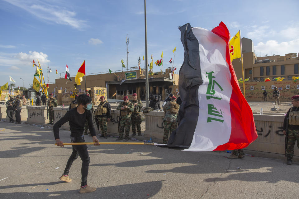 A man waves the Iraqi flag while the Iraqi army soldiers are deployed in front of the U.S. embassy, in Baghdad, Iraq, Wednesday, Jan. 1, 2020. Iran-backed militiamen have withdrawn from the U.S. Embassy compound in Baghdad after two days of clashes with American security forces. (AP Photo/Nasser Nasser)