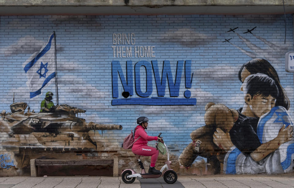 A woman rides a scooter past a graffiti calling for the return of the hostages kidnapped during the Oct. 7 Hamas cross-border attack in Israel, in Kfar Saba, Israel, Monday, Feb. 26, 2024. (AP Photo/Ohad Zwigenberg)