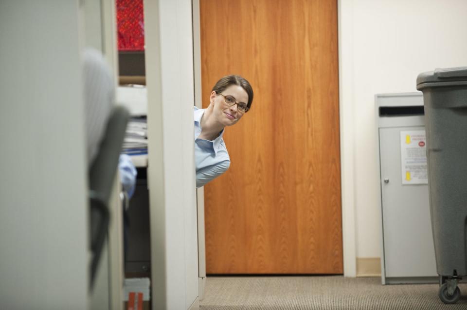 A brunette woman with glasses peeks around a wall of an office and looks at the photographer.